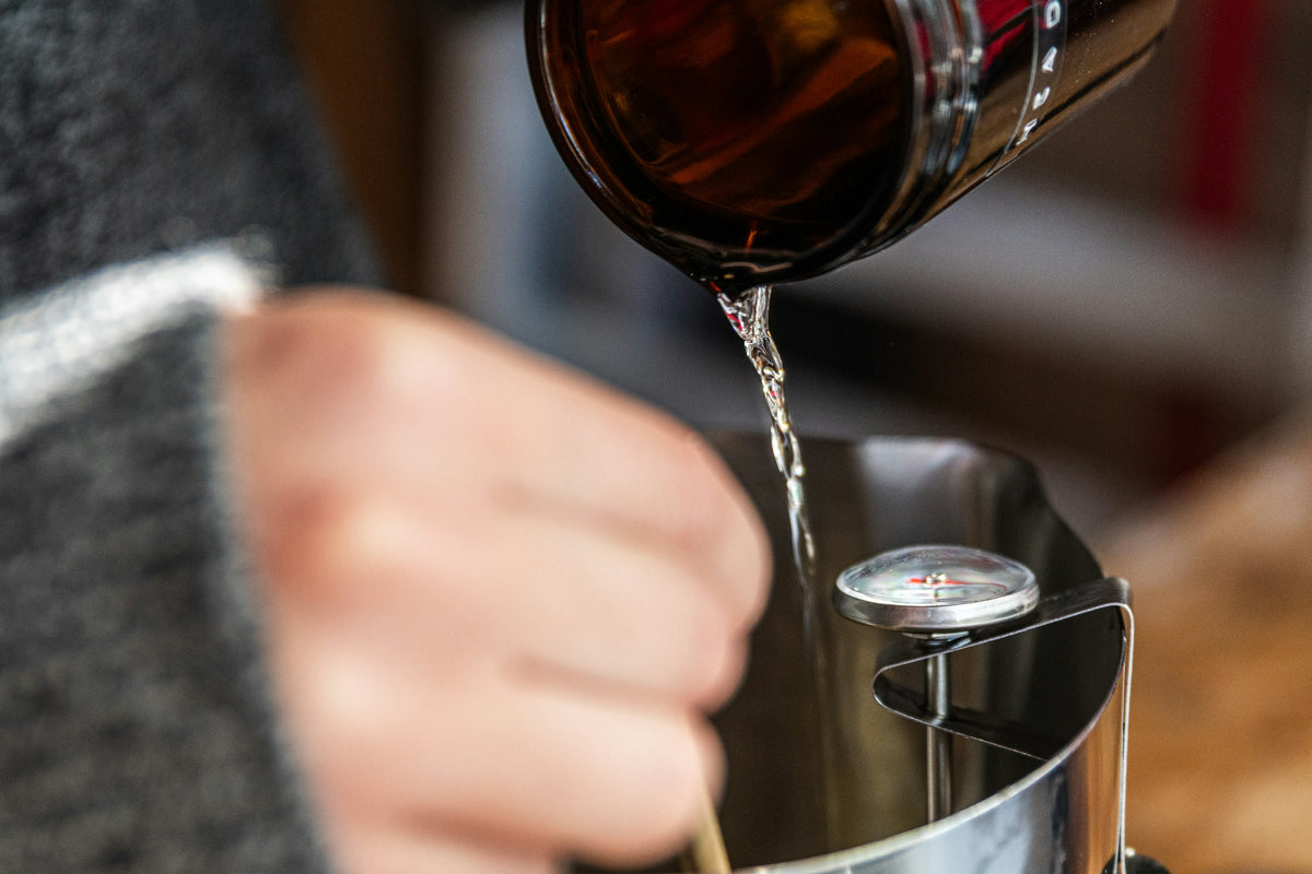Hands pouring melted wax into a metal pitcher with a thermometer